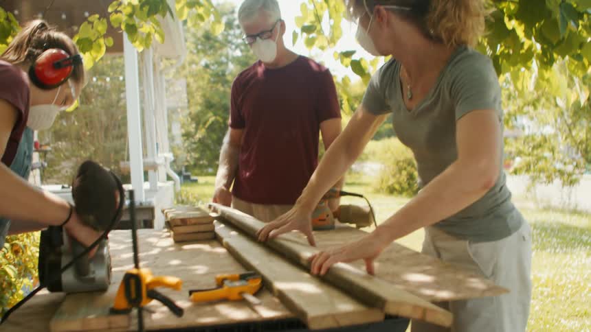 Parents and daughter wearing protective masks using a saw machine and cutting wood in the backyard