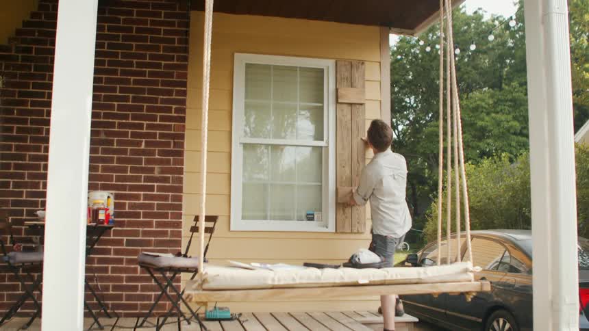 A person in a lightcolored shirt attaches a wooden shutter to a yellowsided house next to a porch swing and a parked car