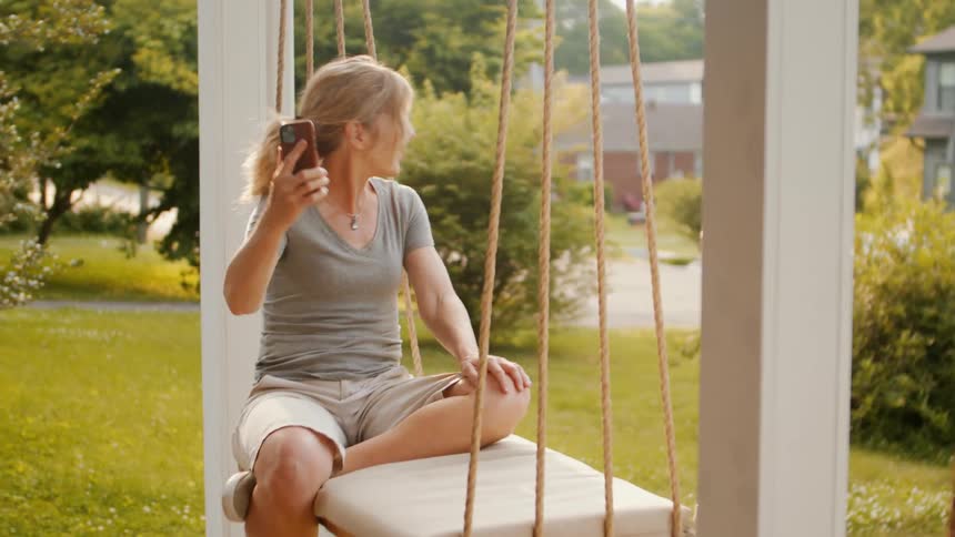 A smiling woman sits on a porch swing happily using her smartphone