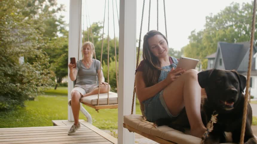 Two people sit on porch swings with a dog one looking at a tablet and the other holding a phone
