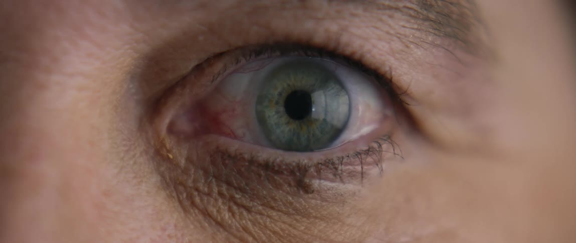 A persons left eye with a green lens blinking slowly with wrinkles visible around the eyelid