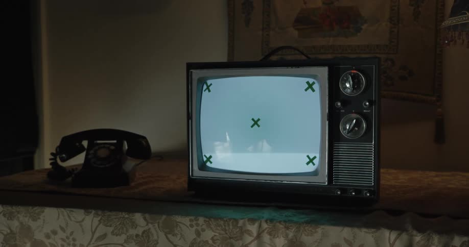 An old television displaying a blank screen with green x marks in the corners and center sits on a table next to a rotary dial telephone