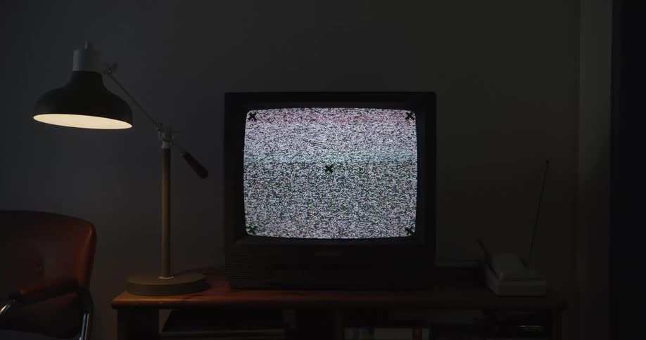A vintage television on a wooden table emits static accompanied by a telephone and a desk lamp in a dimly lit room