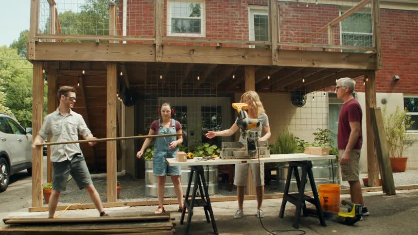 Family in the backyard of a house with one of them placing a piece of wood on a table with a saw