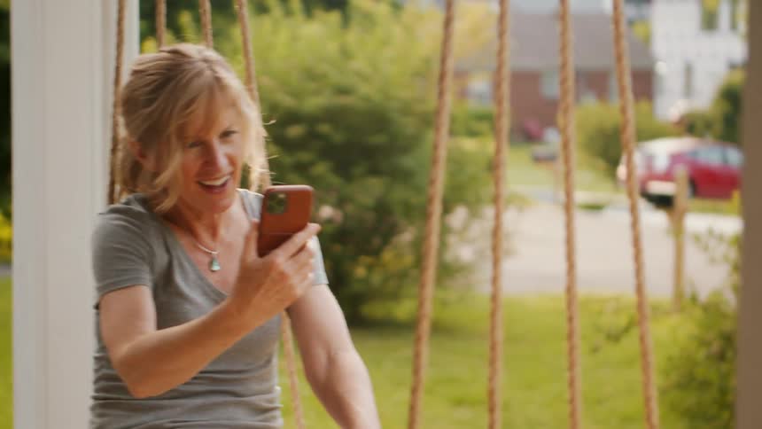 A woman sits on a porch swing while video calling on her smartphone