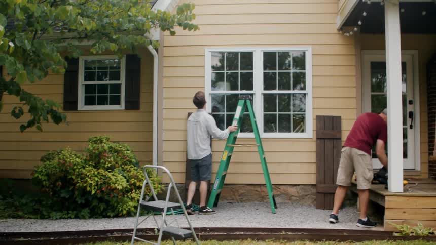 Two men in the backyard of a house with one of them handling a green ladder