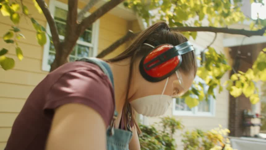 A young woman wearing a face mask and headphones sanding a plank with a power tool outdoors