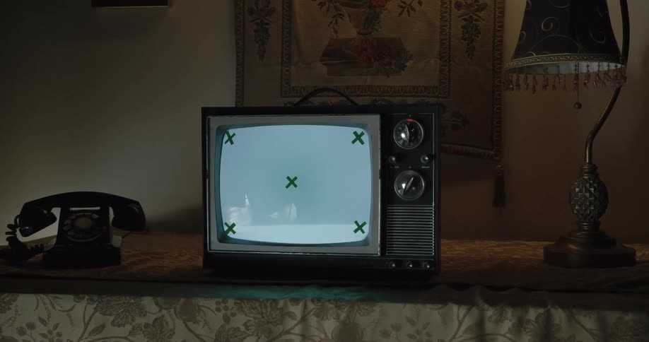 The camera pans right and moves closer to an old portable crt tv showing a flickering blank screen with tracking markers in a dimly lit vintage room