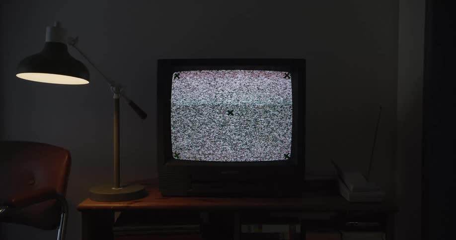 Old school tv with white noise flashing on the screen sitting on a small table next to low lit lamp