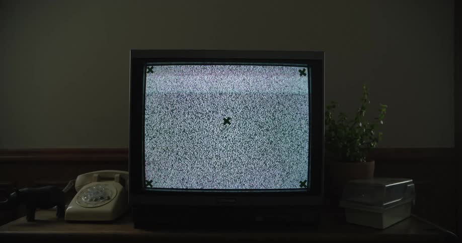 A vintage crt television displays static with tracking markers sitting on a desk next to a rotary phone and plant in a dim room