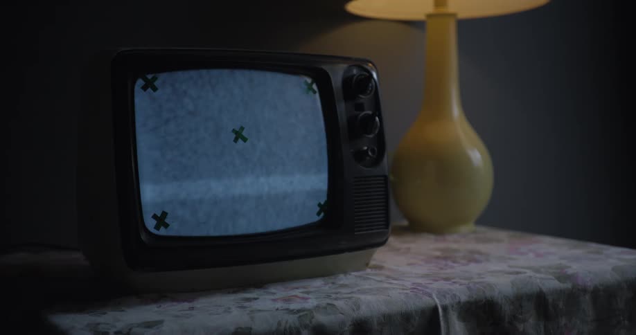 An old television on a floral tablecloth displays static with green x marks on the screen beside a yellow lamp
