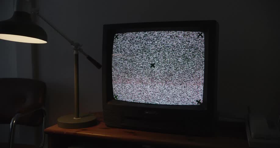 A vintage television displaying static sits on a wooden surface beside a lit desk lamp in a dimly illuminated space