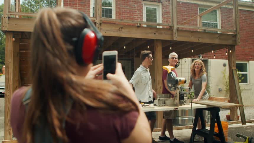 A young lady wearing protective headphones uses her smartphone to take photos of her parents and her husband standing side by side in front of a workbench with a saw on top while woodworking at midday