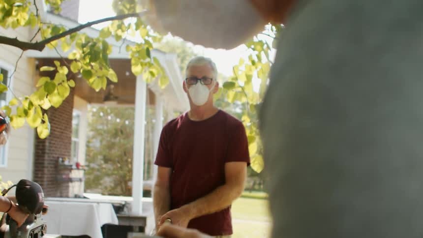 A family work on woodworking outdoors with one sanding a wooden plank while another uses a saw in the background