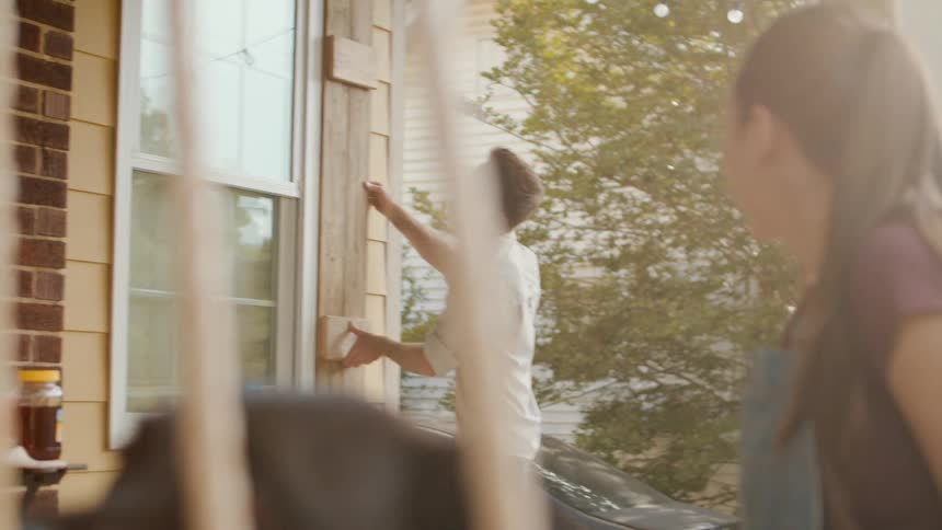 A person installs wooden shutters on the exterior of a house while another watches nearby from a porch swing