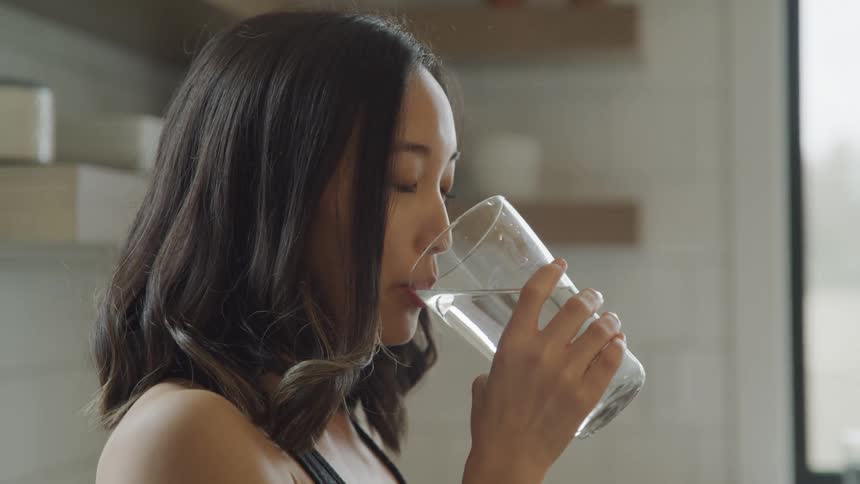 A woman drinks a large glass of water standing in a kitchen