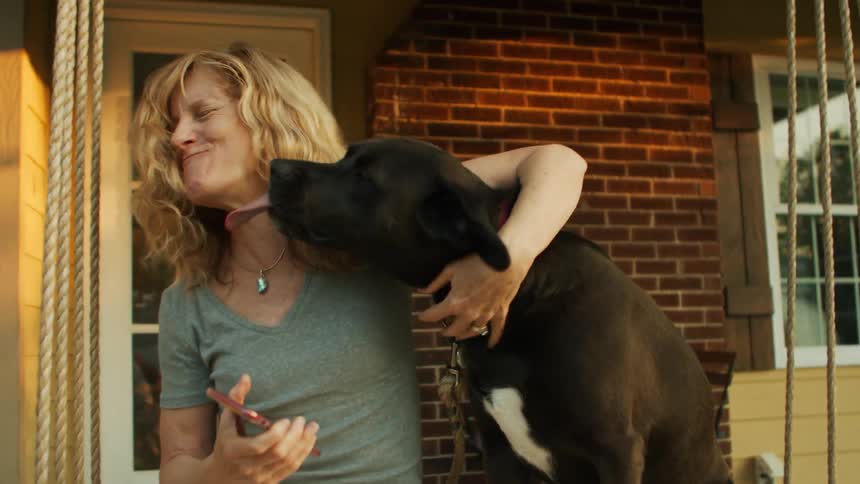 A woman wearing a gray shirt holds a phone and hugs a large black dog with a white patch on its chest in front of a house with brick walls and a door