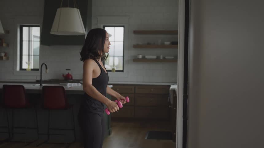 A young woman lifts dumbbells while exercising in her kitchen at midday
