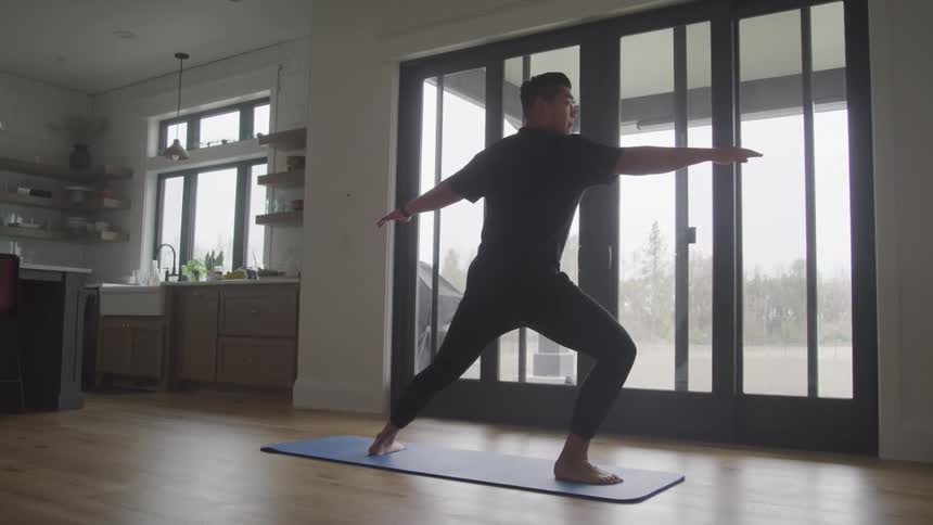 A person performs a yoga pose on a blue mat in a modern kitchen with large windows and wooden floors