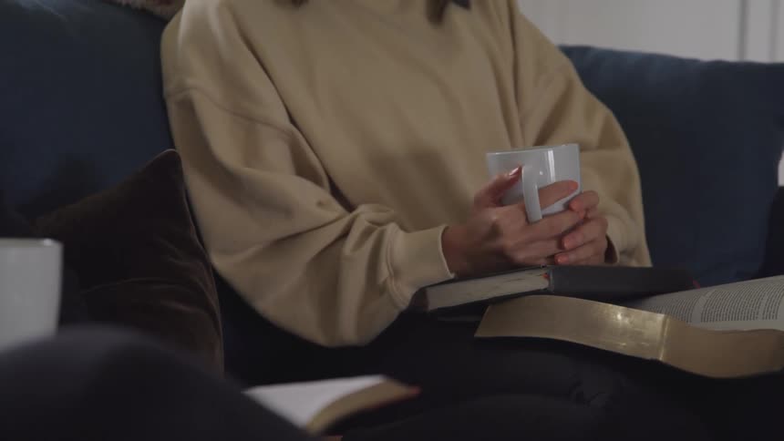 A person wearing a beige sweater sits on a couch holding a white mug and an open book
