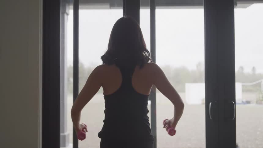 A person stands extending her arms holding small red weights in a sunlit room with large glass doors