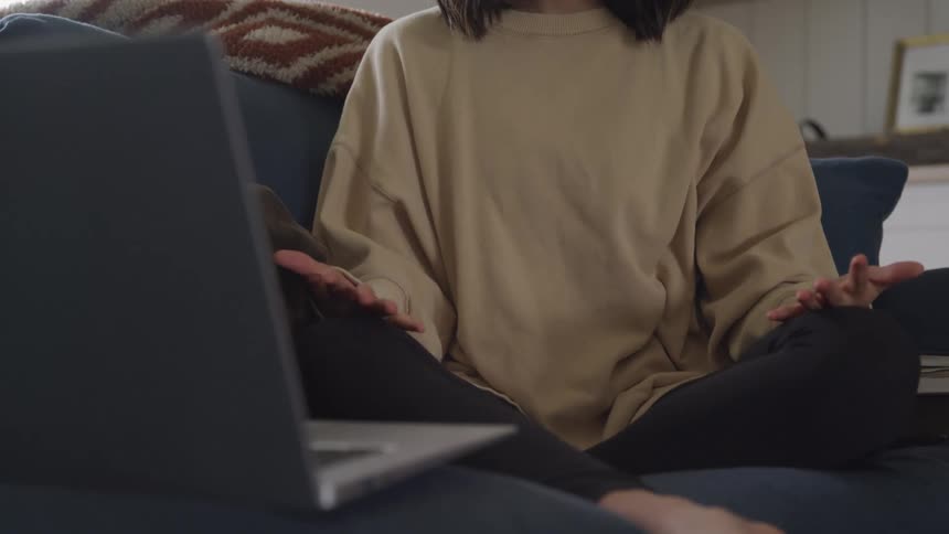 A person sits cross legged on a couch wearing a beige sweatshirt with hands resting on the knees and a laptop open in front