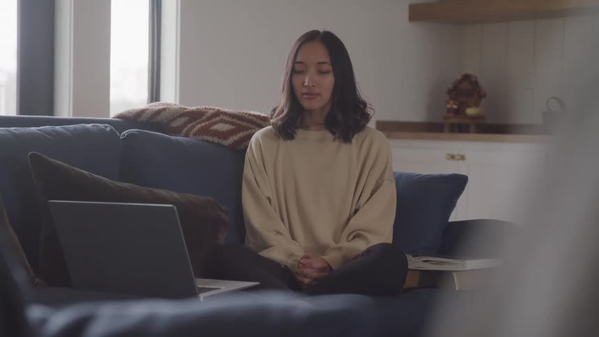 A woman is meditating on the couch near her laptop