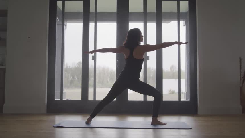 Woman takes a yoga pose while standing on a mat near a glass door showing a scenic landscape