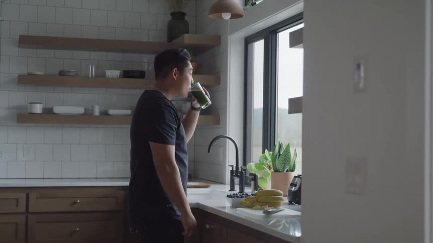 A man drinks green liquid from a glass cup while standing close to the kitchen window at dawn