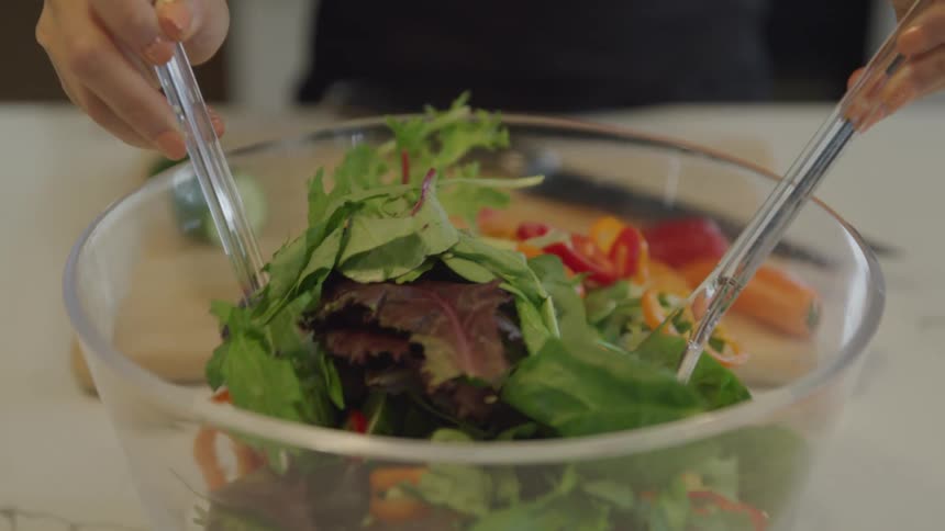 Tossing a mixed green salad in a small clear glass bowl on counter