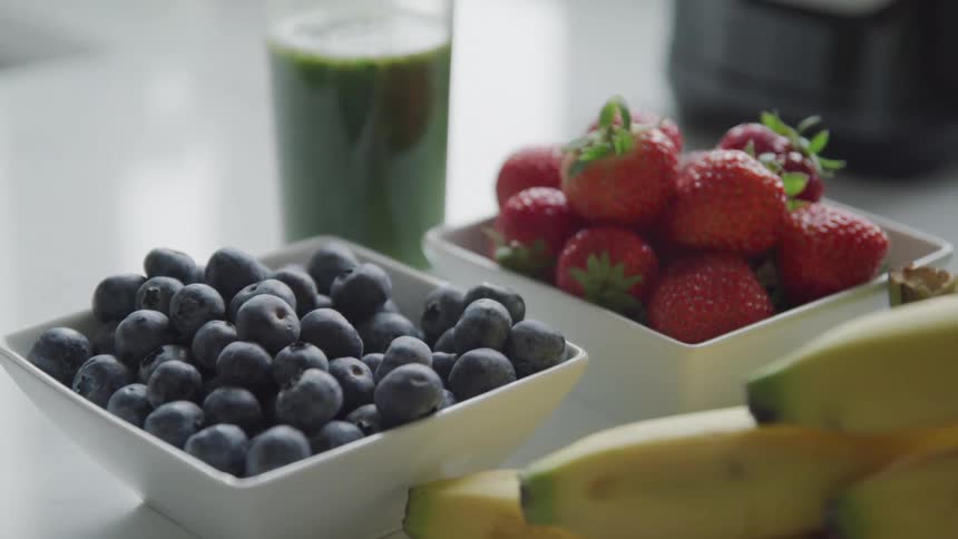 A table holds bowls of ripe blueberries and strawberries a bunch of bananas and a glass filled with a vibrant green smoothie