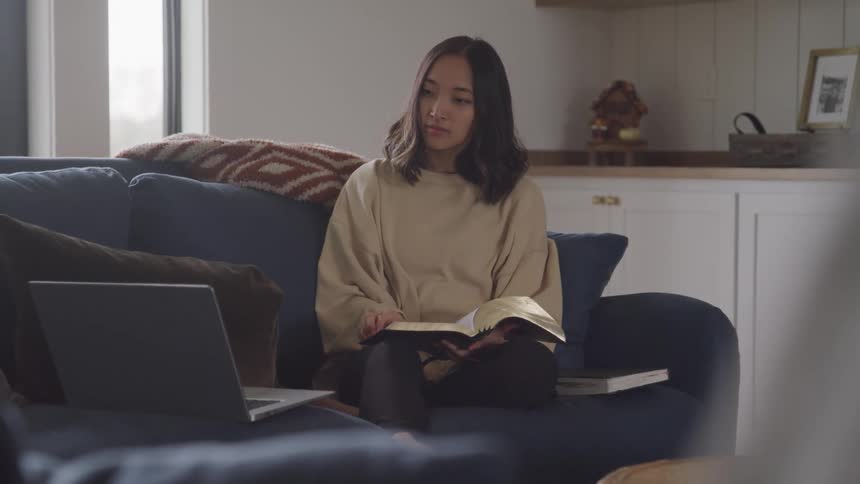 A woman sits on the couch flipping through a book while her laptop is nearby