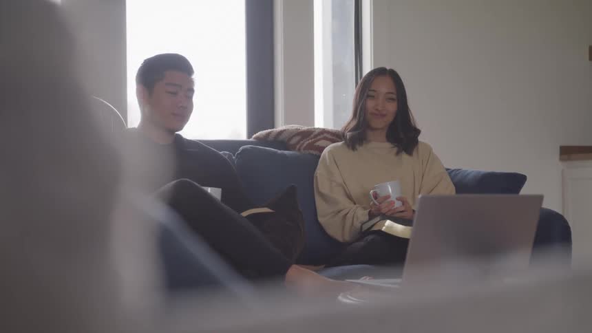 A man and woman are sitting on a couch with coffee cups in front of a laptop on a table