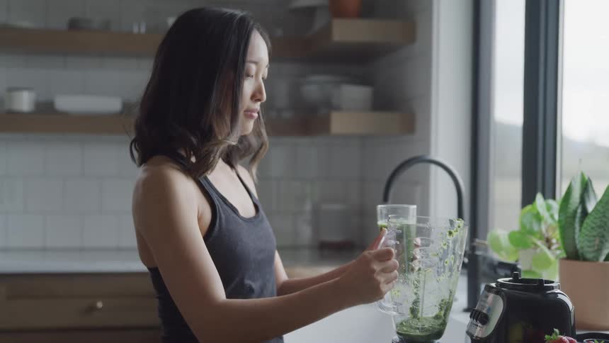 A woman prepares and drinks a green vegetable juice while standing in a kitchen