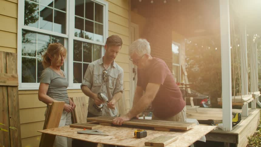 A family works together on a woodworking project outdoors gathered around a large wooden table near their house