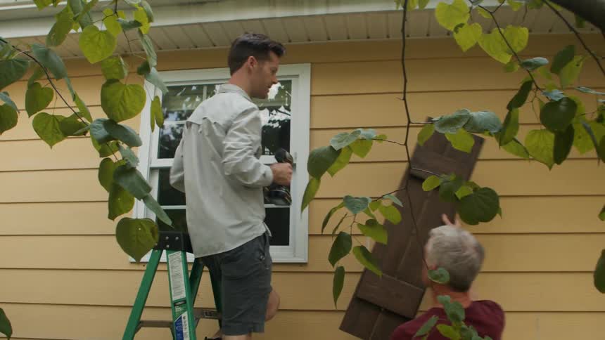 Two people install a wooden shutter beside a window on the exterior of a yellow house with one standing on a green ladder holding a drill and the other assisting from the ground