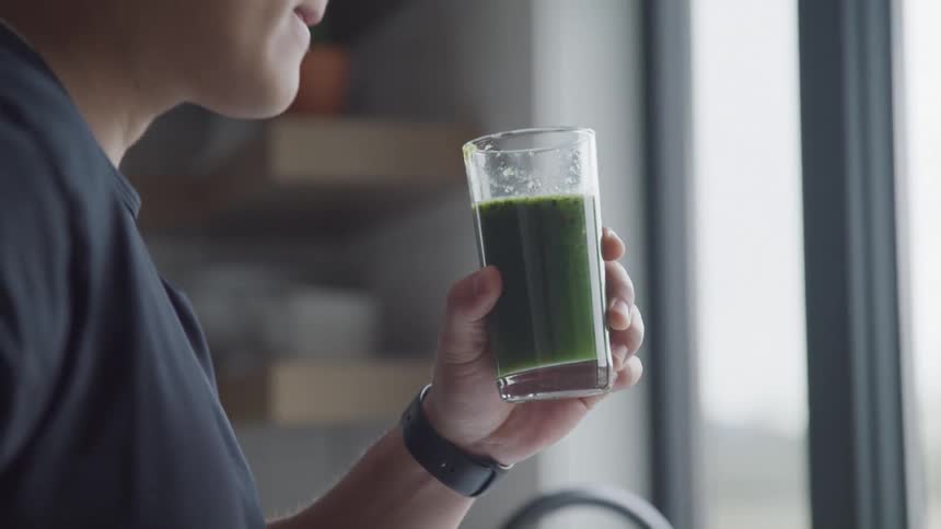 A young asian man drinks green juice from a glass while standing in his kitchen at midday