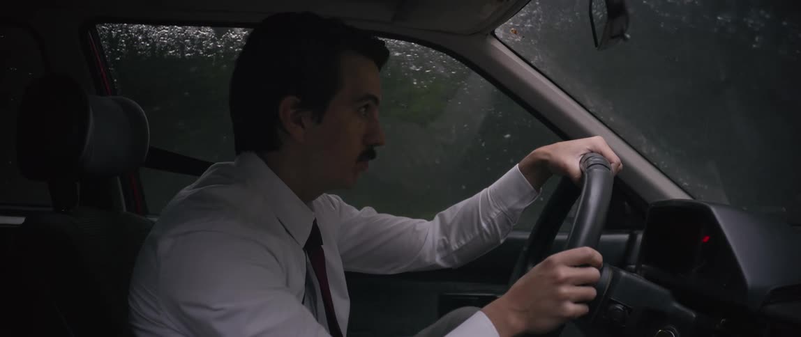A man is shown driving a car in a dimly lit environment and rainy conditions