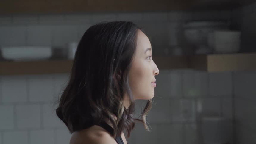An asian woman is gazing out the window while standing in her kitchen