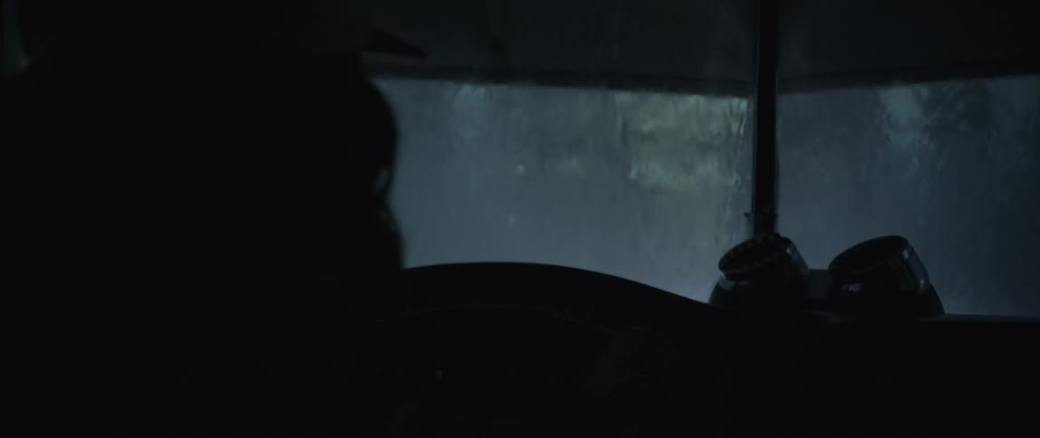 A shadowed figure is faintly visible inside a vehicle with a distorted view of a rainsoaked dim environment through the windshield