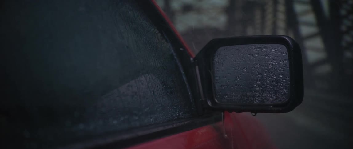 In the rain a close up focuses on the water beaded side mirror and window of a red car as it drives over a bridge
