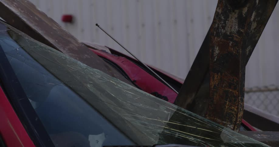 A closeup shows the cracked windshield of a red car as heavy machinery lowers onto it in what appears to be a junkyard