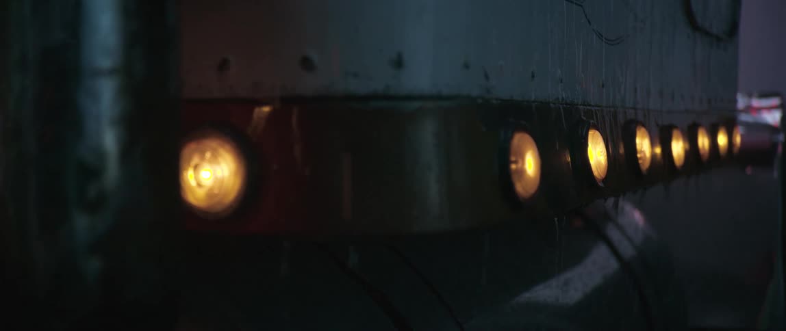 A closeup shot shows the rear of a semitruck at night illuminated by its tail lights and side marker lights with a partially visible exhaust pipe under heavy rain