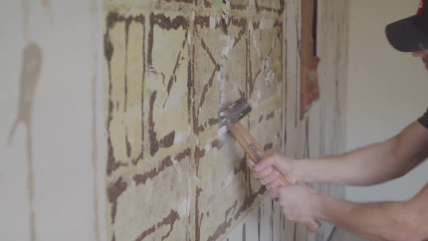 A person uses a hammer to chip away old plaster from a weathered wall