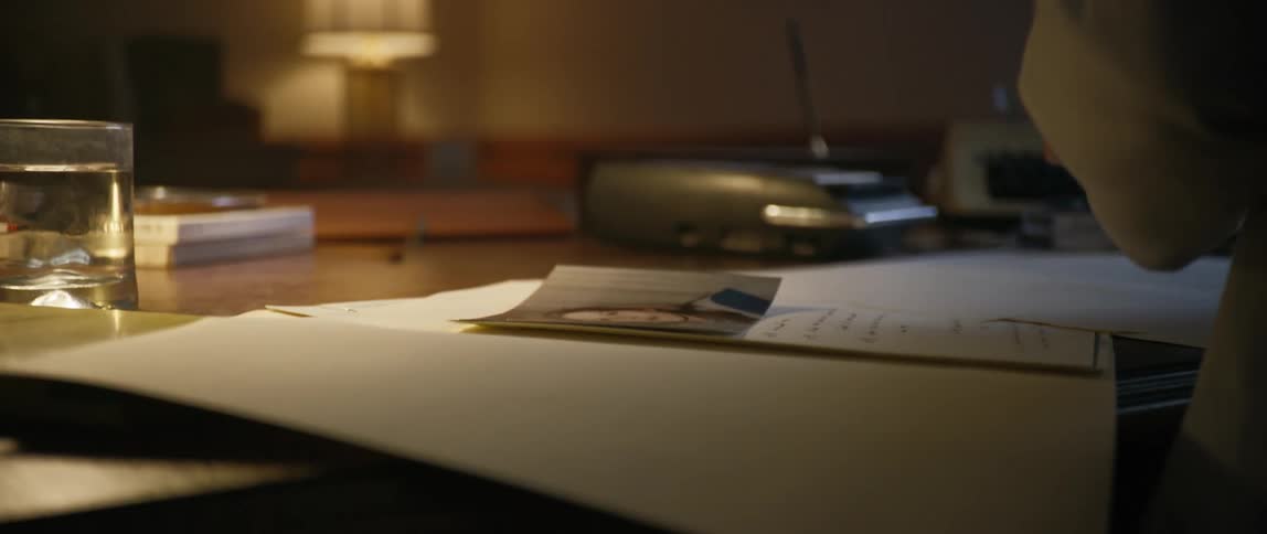 A man in a lab coat picks up vhs tapes from a desk in a dimly lit office