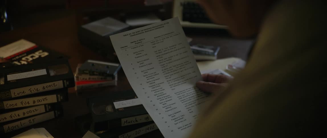 Man reviews lists while sitting at a desk stacked with audio and vhs tapes