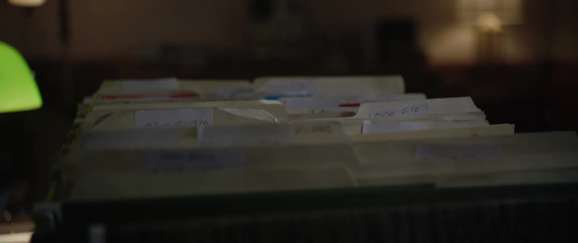 A dimly lit room features a stack of manila file folders on a desk