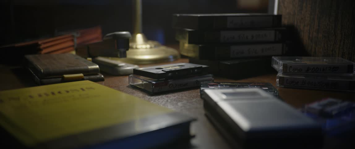 A cassette tape lies among a yellow book a cassette recorder vhs tapes and a vintage lamp all scattered on a wooden surface in a dimly lit room