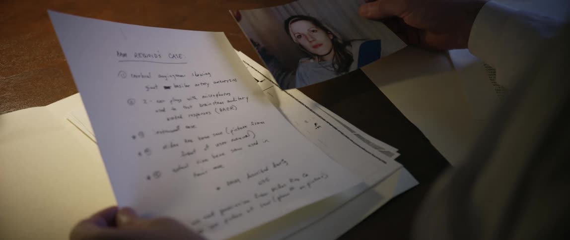 A doctor seated at a desk examines a file containing a document with a womans photograph