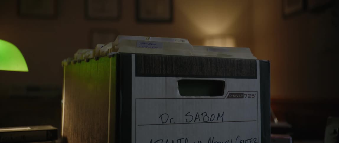 A box filled with files sits on a wooden desk surrounded by cassette tapes and a green desk lamp in a dimly lit room